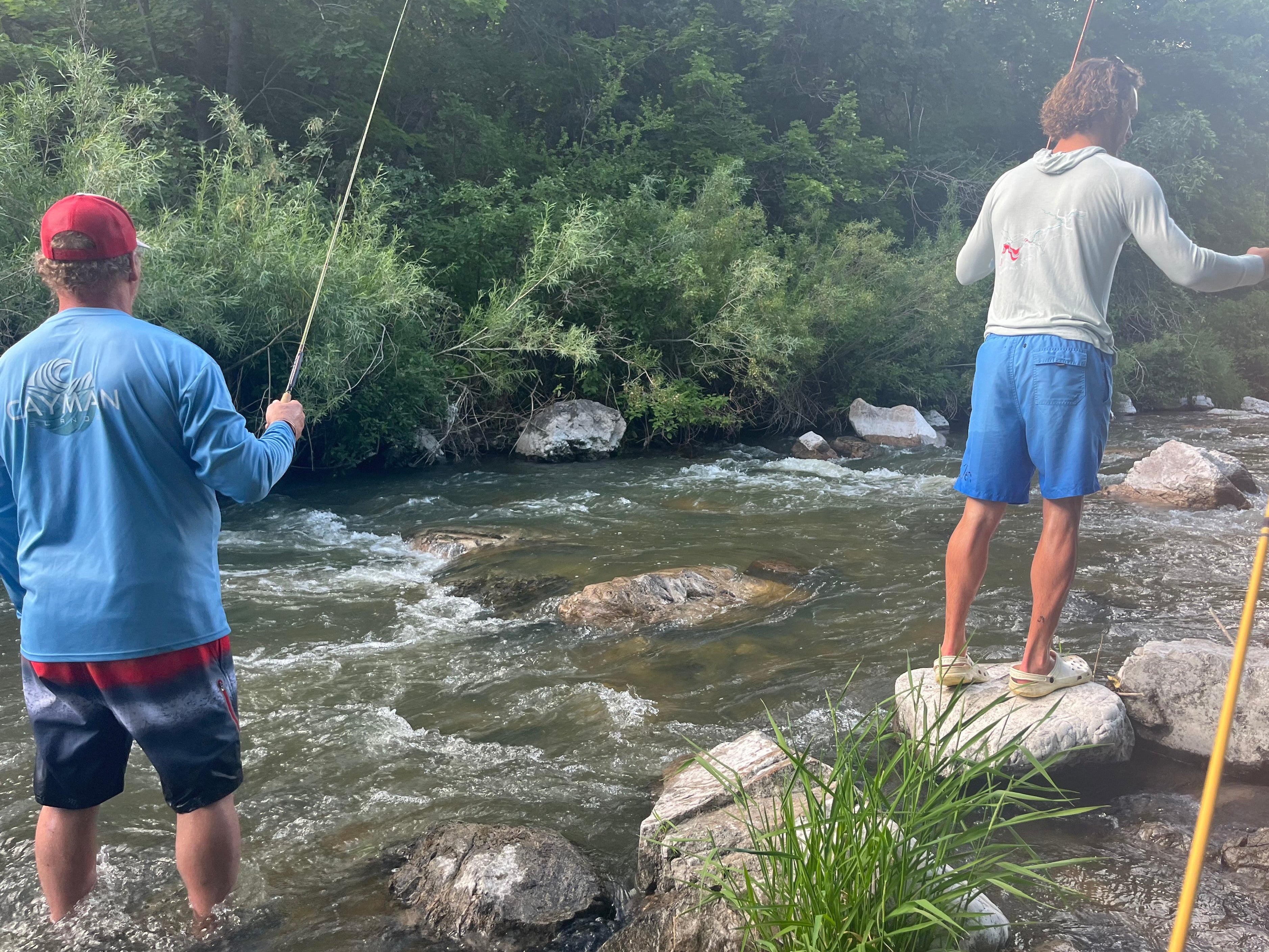 Two people fishing in a river with rocks and greenery around.