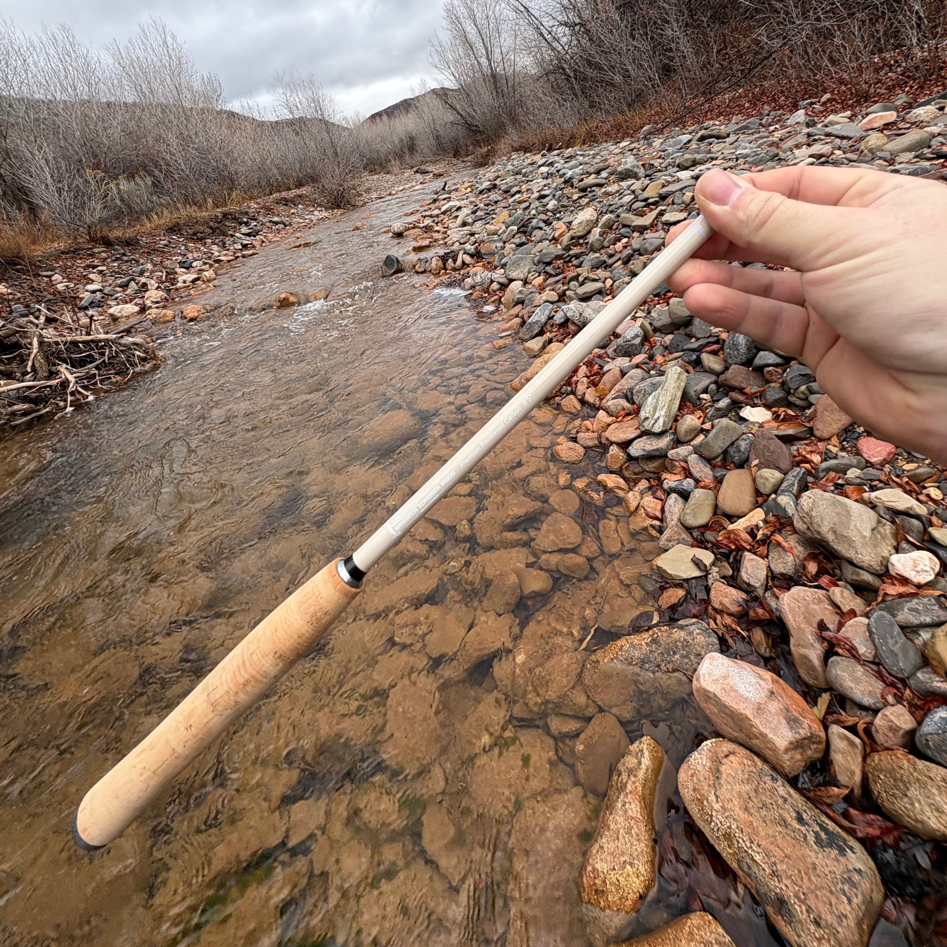 Fishing rod held by a hand over a stream with stones and water.