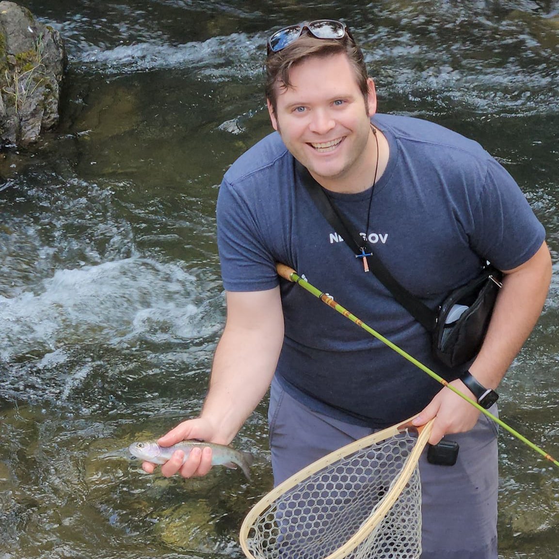 Smiling man holding a small trout and net while fishing in a rocky mountain stream using a green Wasatch Tenkara Rods Baby RodZilla rod, showcasing an ideal backcountry fly fishing moment.