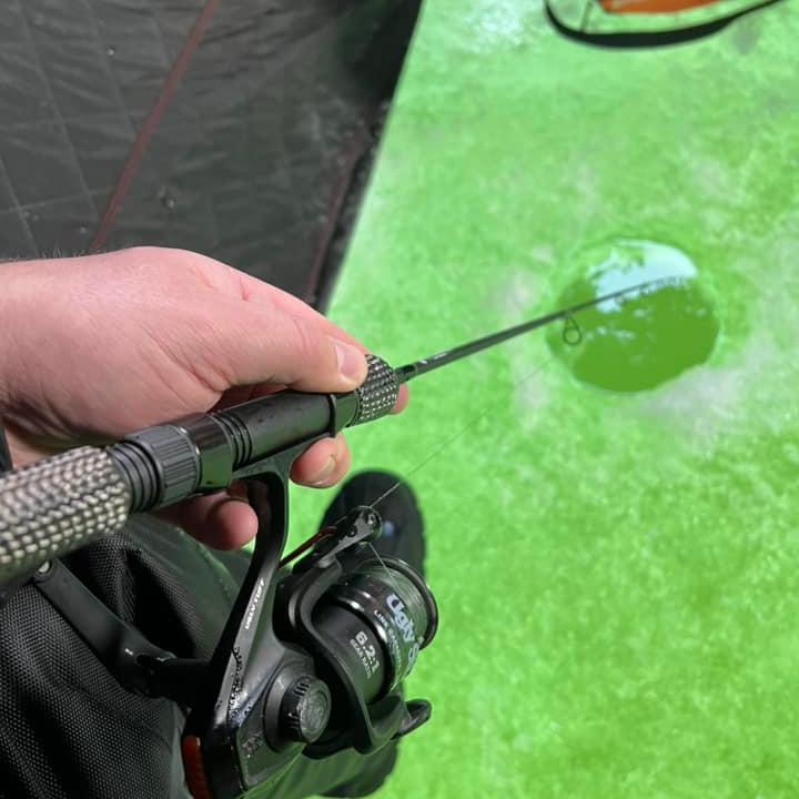 A hand holding a Wasatch Tenkara Rods - Slot Buster rod and reel, poised over a green ice fishing hole inside an ice shelter, ready for the next catch.