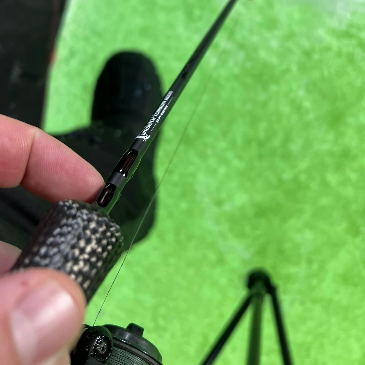 A close-up shot of a hand holding the textured handle of a Wasatch Tenkara Rods - Slot Buster ice fishing rod, highlighting the rod's sleek design and branding, with a green ice fishing surface in the background.