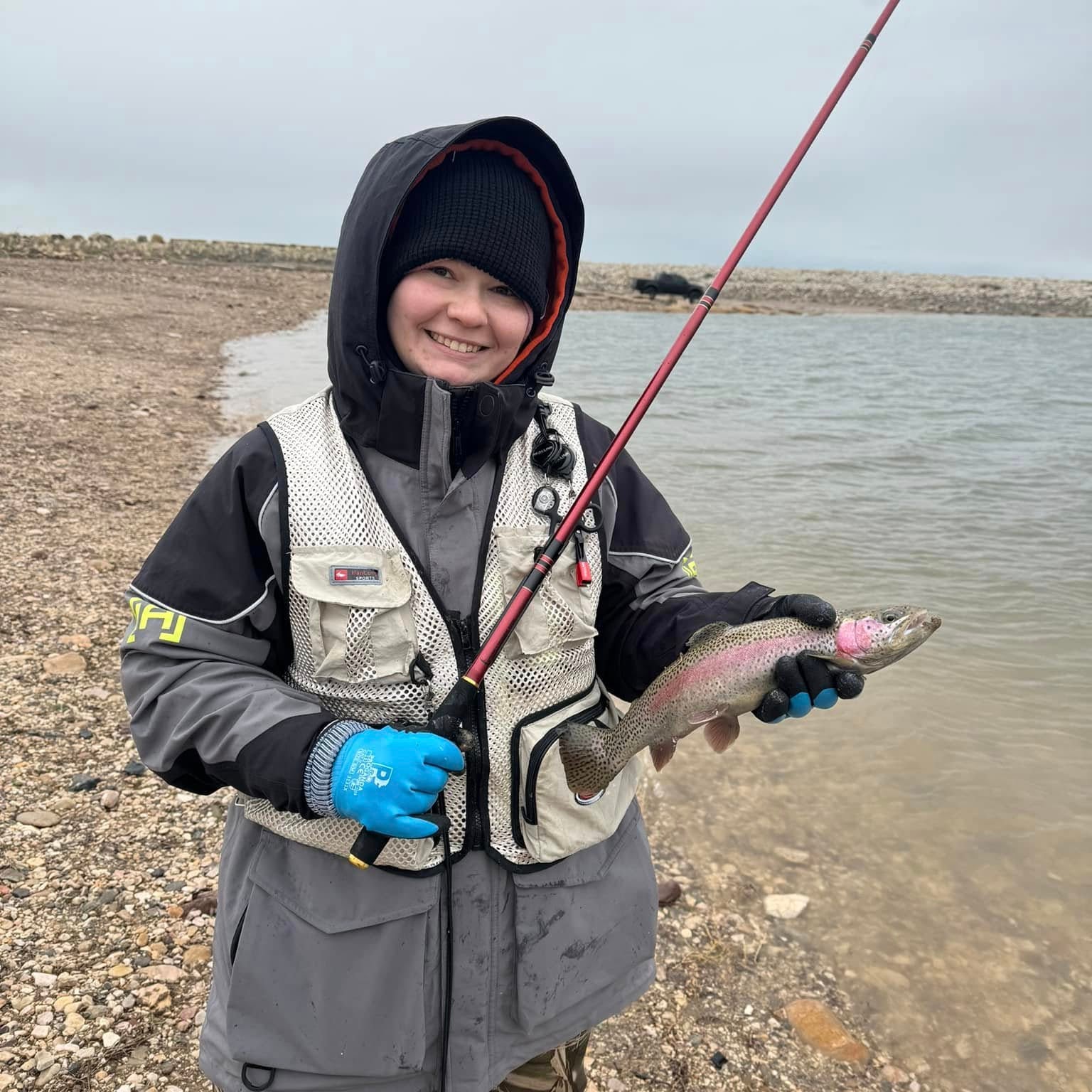 Angler bundled in winter gear smiles while holding a freshly caught rainbow trout along a rocky shoreline. The red Akai Samurai Tenkara Rod is visible, showcasing a successful cold-weather catch.