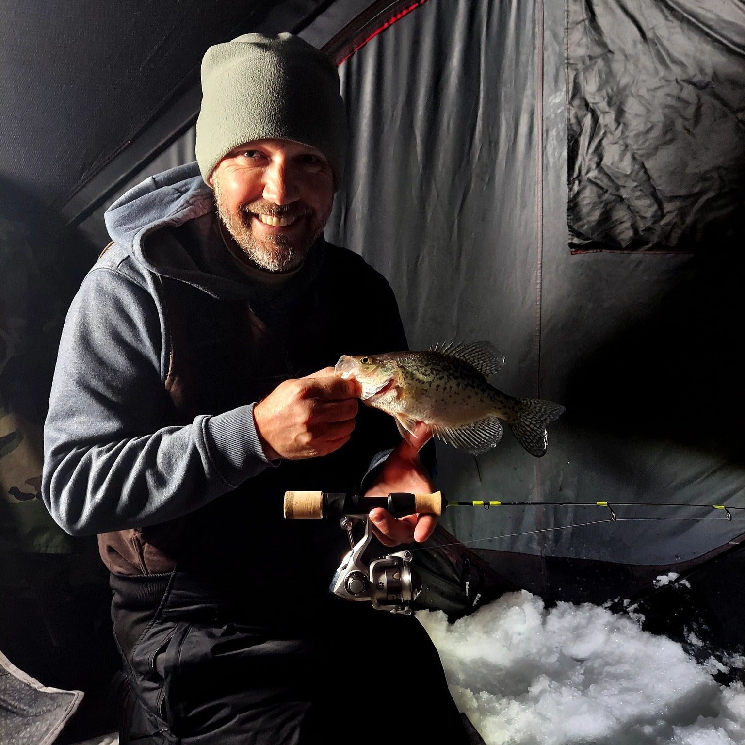 A smiling angler dressed warmly in a beanie and hoodie holding a crappie fish and a Wasatch Tenkara Rods - Ice Breaker ice fishing rod inside an ice fishing shelter, with snow and gear visible in the background.