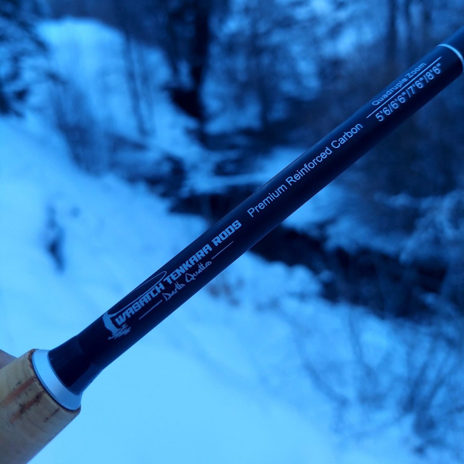 A close-up of a Wasatch Tenkara Rods - Darth Quattro fishing rod labeled "Premium Reinforced Carbon" and "Quadruple Zoom" with length options of 5'6"/6'6"/7'6"/8'6". The background features a snowy, forested stream, captured in a cool blue tone that reflects the chilly winter environment.