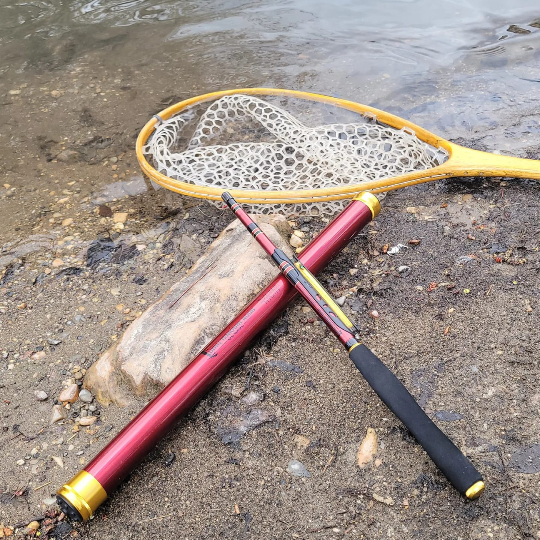 Akai Samurai Tenkara Rod placed on the sandy riverbank next to its matching red rod tube and a wooden landing net with rubber mesh, capturing a scenic and ready-to-fish moment by the water's edge.