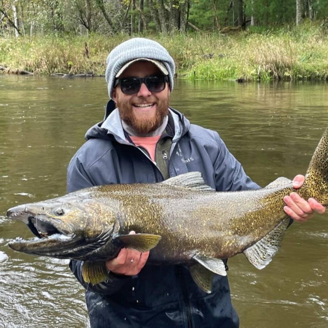 Smiling angler standing in a river, holding a massive Chinook salmon with both hands, wearing a beanie, sunglasses, and a waterproof Simms jacket—caught using Wasatch Tenkara Rods gear.