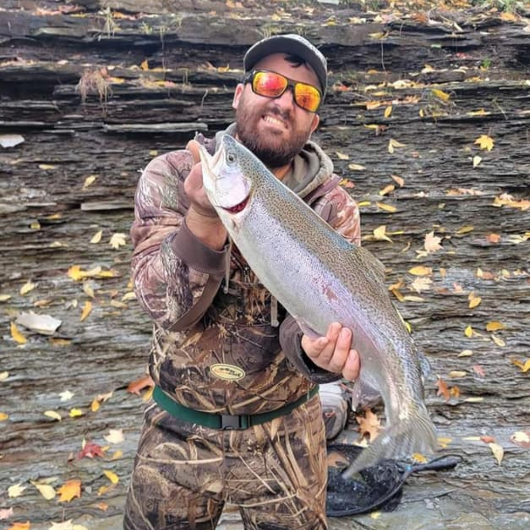 Smiling angler in camouflage waders holding a large rainbow trout in a rocky, leaf-covered streambed—showcasing the capability of Wasatch Tenkara Rods for landing big fish in rugged environments.