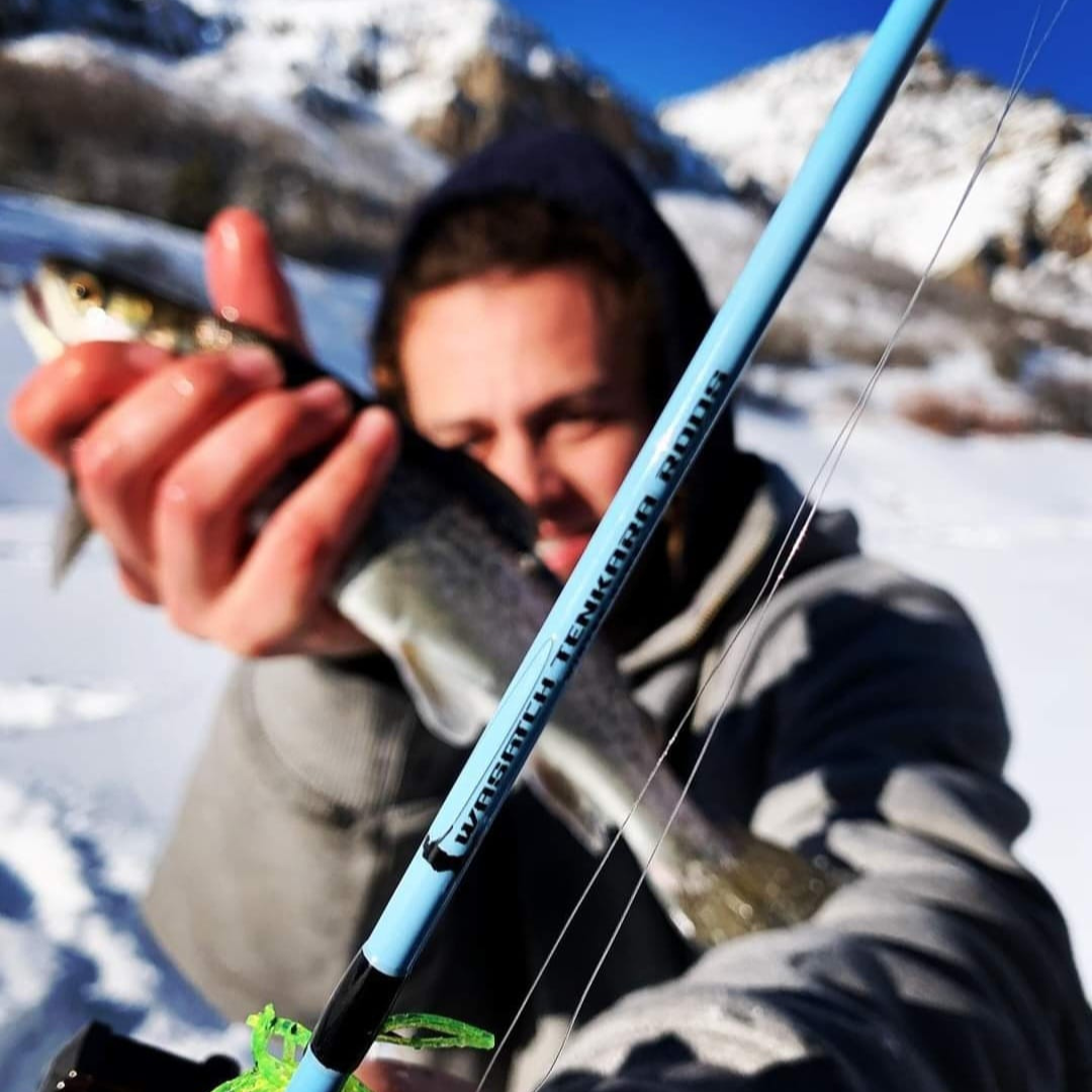 A person holding a freshly caught fish in a snowy mountain setting, with a close-up of the light blue Wasatch Tenkara Rods - Winterhawk ice rod in the foreground, showcasing the rod's branding.