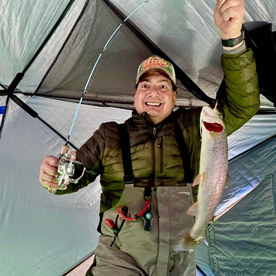 A smiling angler inside an ice fishing shelter holding a Wasatch Tenkara Rods - Winterhawk ice fishing rod in one hand and a freshly caught trout in the other, showcasing a successful and joyful fishing experience.