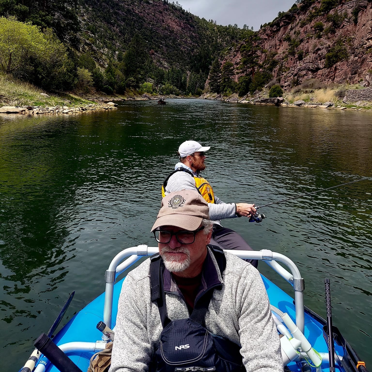 Two anglers fishing from a blue inflatable raft on a scenic river, surrounded by pine-covered cliffs and clear water, using the Wasatch Tenkara Rods Command Force spin rod—ideal for wilderness fishing and rugged performance.