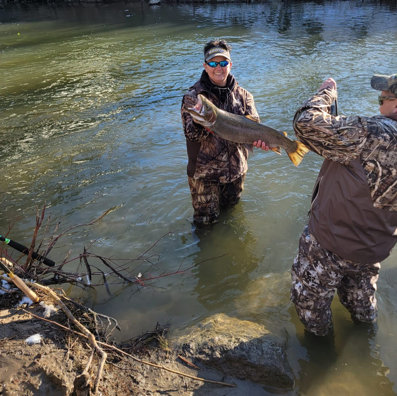 Smiling angler in camo waders holding a massive trout in shallow river water while another person snaps a photo, with a green Wasatch Tenkara Rods SIYCO rod resting on the bank—showcasing the rod’s capability for landing big fish.