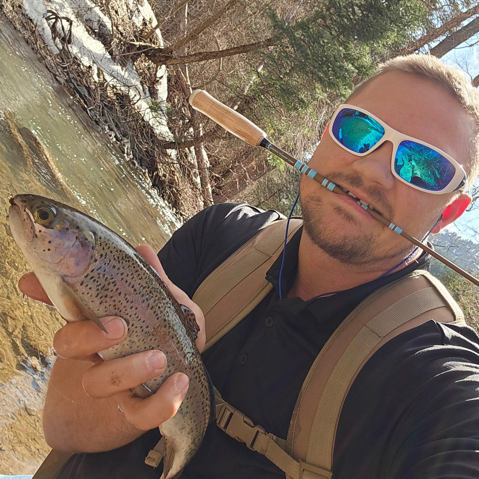 Fly fisherman holding a rainbow trout beside a clear mountain stream, with a Wasatch Tenkara Snub Nose IV rod in his mouth, showcasing ultralight gear for backcountry fishing adventures.