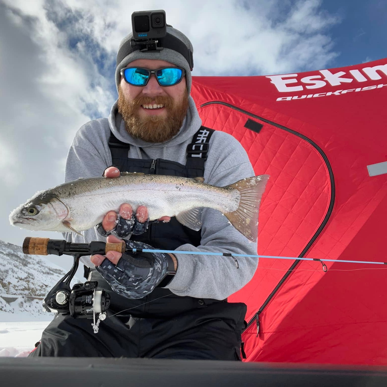 A smiling ice fisherman wearing a GoPro and sunglasses holds a freshly caught trout alongside a Wasatch Tenkara Rods - Winterhawk ice fishing rod, with a red Eskimo ice fishing shelter and snowy mountain backdrop.
