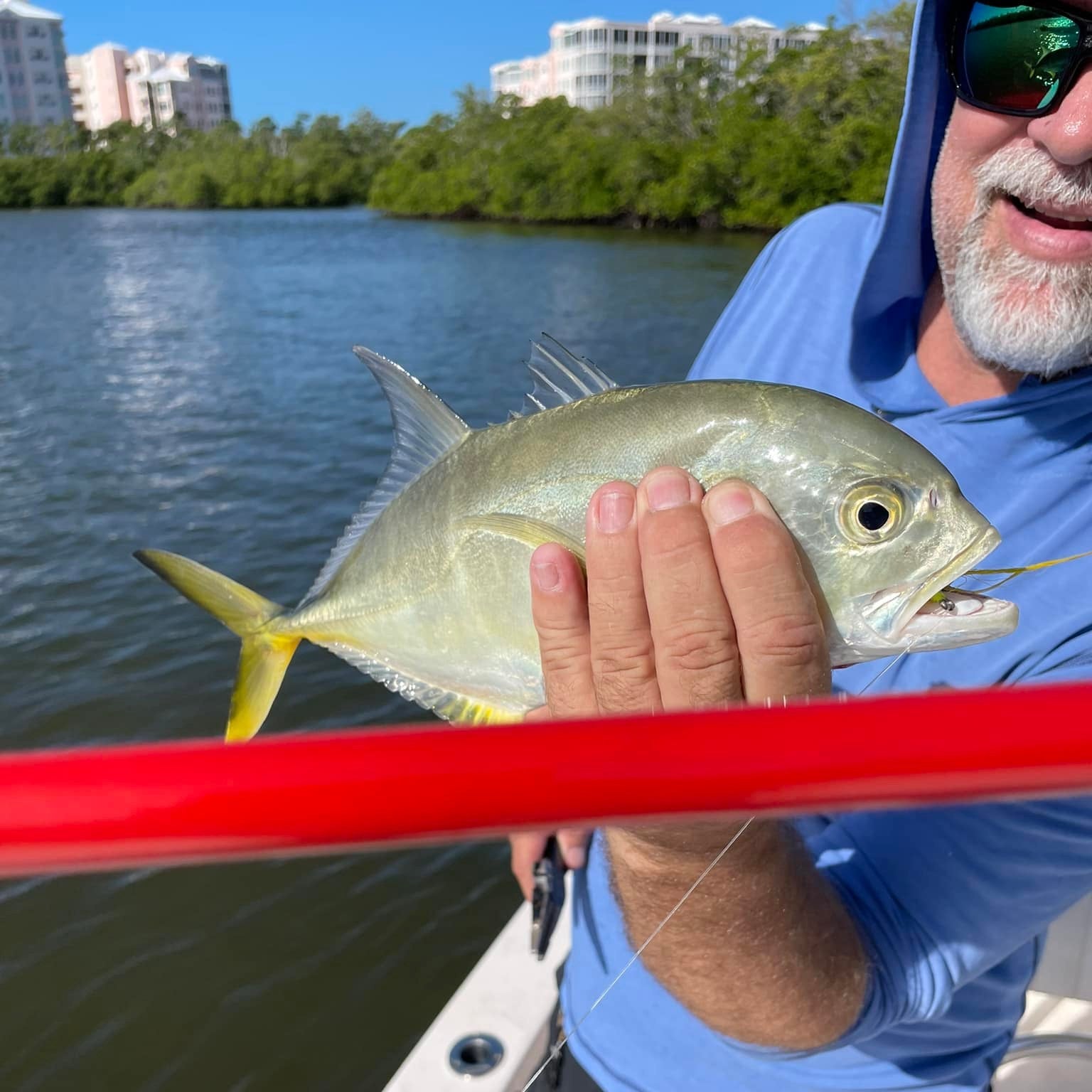Angler holding a bright silver jack fish caught with a Wasatch Tenkara Rods Phoenix Rising tenkara rod in coastal saltwater, with waterfront buildings and mangroves in the background.