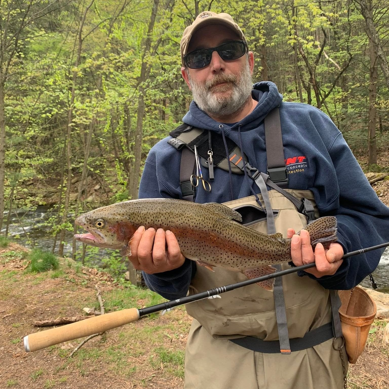 A Wasatch Tenkara Rods fishing rod, the Darth Quattro model, is held upright in front of a large, vibrant rainbow trout caught on the line. The fish, displaying bright pink and greenish hues, is clearly visible and quite large, while the rod itself is dark and sleek, set against a natural outdoor background of trees and a river.